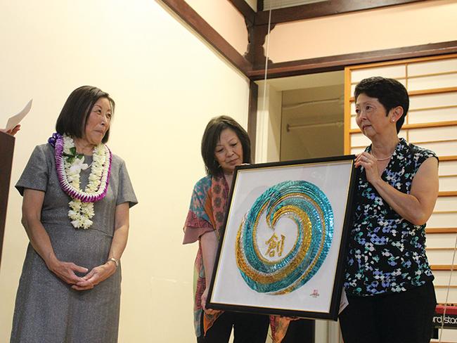 Two woman present a framed piece of origami artwork to a third woman.